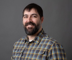 professor wearing a dark blue and green plaid button up shirt in front of a gray backdrop